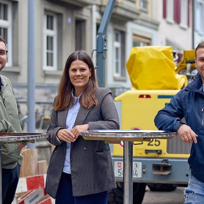 Zwei Männer und eine Frau an Stehtischen vor einem Baustellenfahrzeug.
