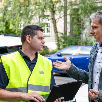 Zwei Männer stehen in einer grünen Quartierstrasse vor einer Ladestation mit IWB-Logo.