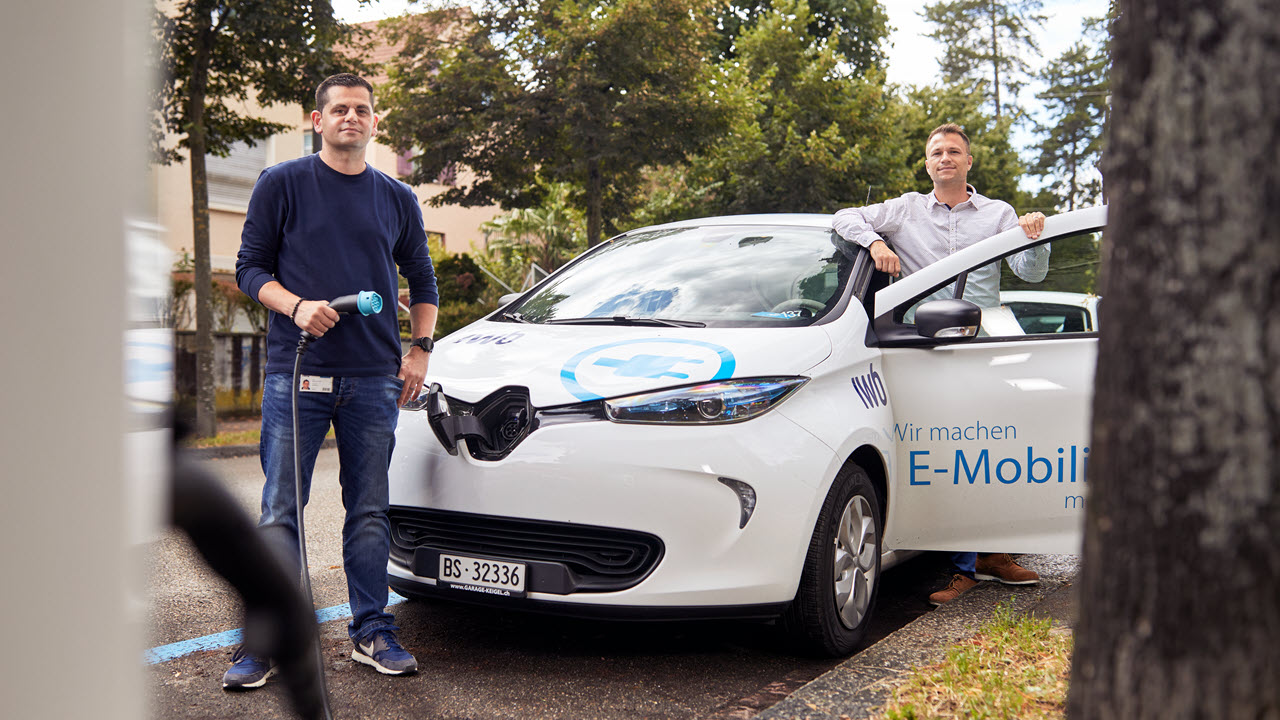 Zwei Männer stehen vor einem Elektroauto, das am Strassenrand geparkt ist. Einer hält ein Ladekabel in der Hand vor der Ladestation, der andere steht beim Fahrzeug.