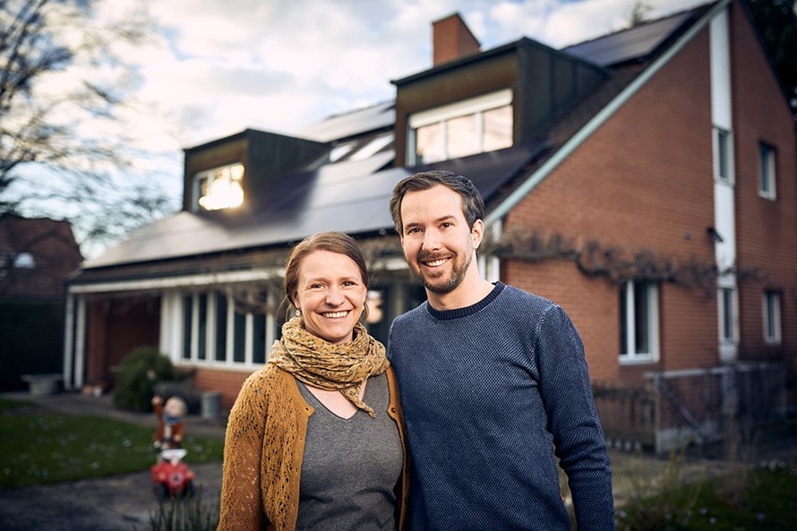 Eine Frau und ein Mann stehen Arm in Arm vor einem Einfamilienhaus mit Solaranlage auf dem Dach.