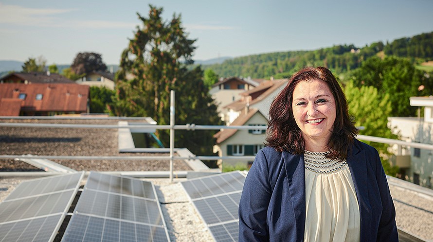 Porträt einer Frau auf dem Dach eines Mehrfamilienhauses. Im Hintergrund ist eine Solaranlage zu sehen und weiter hinten die Jurahügel.