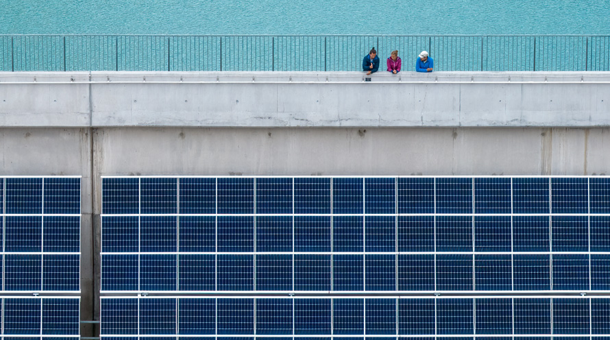 Drei Personen stehen auf einer Staumauer. Unterhalb hängen grosse Solarpanels.
