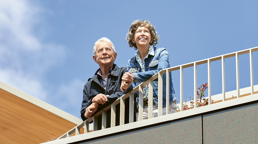 Ein älteres Ehepaar lehnt über ein Balkongeländer und blickt in die Ferne.