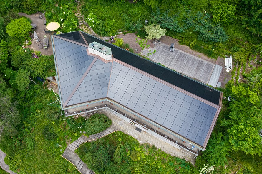 Ein Haus inmitten von Grün von oben. Die ganze, geknickte Dachfläche ist mit Solarpanels bedeckt.