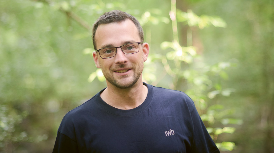 Janik aus den Erlen, Forstwart, IWB. Porträt eines Mannes im T-Shirt, der inmitten von Grün im Wald steht.