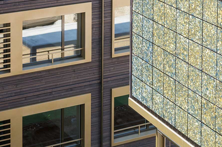 Detailansicht eines Neubaus mit Holzfassade. Im Vordergrund ist ein Balkon zu sehen, dessen Brüstung mit farbigen Solarpanels bedeckt ist.