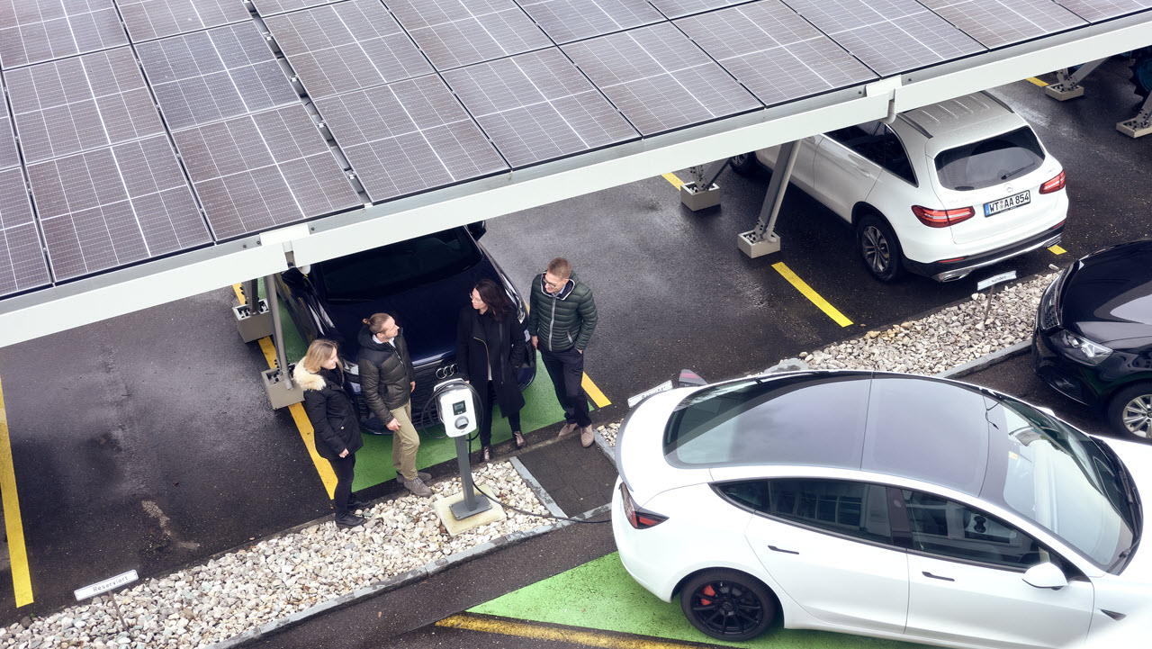 Eine Frau und ein Mann stehen unter einem Carport, das mit Photovoltaik-Panels bestückt ist. Rund herum sind Autos parkiert.