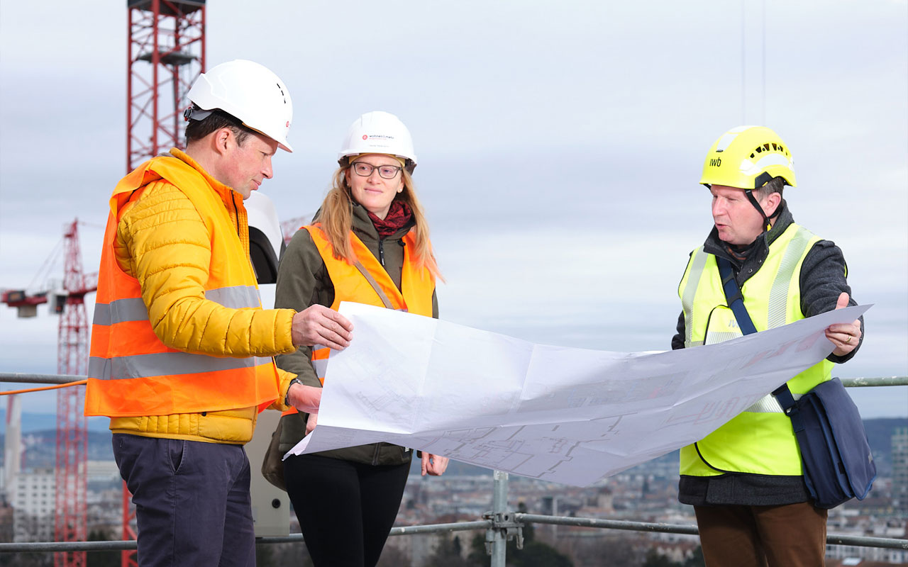 Eine Frau und zwei Männer stehen auf dem Dach einer Baustelle. Sie blicken auf einen grossen Plan. Im Hintergrund steht ein roter Kran.