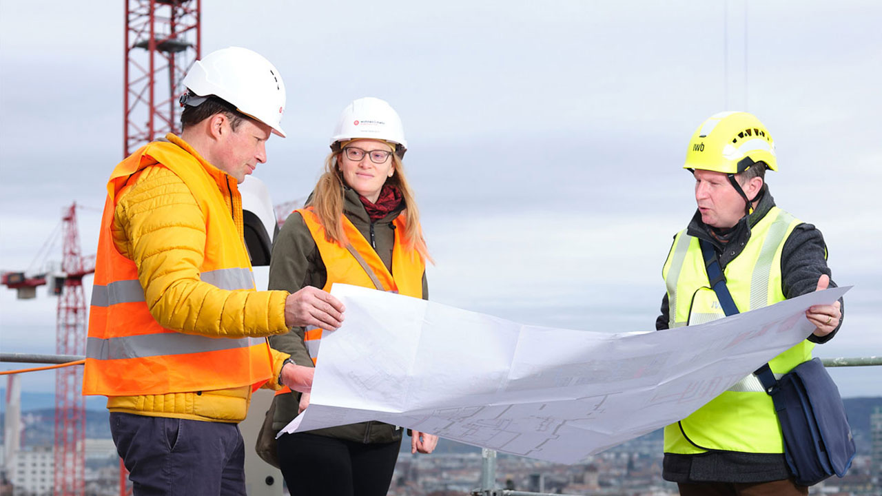 Eine Frau und zwei Männer stehen auf dem Dach einer Baustelle. Sie blicken auf einen grossen Plan. Im Hintergrund steht ein roter Kran.