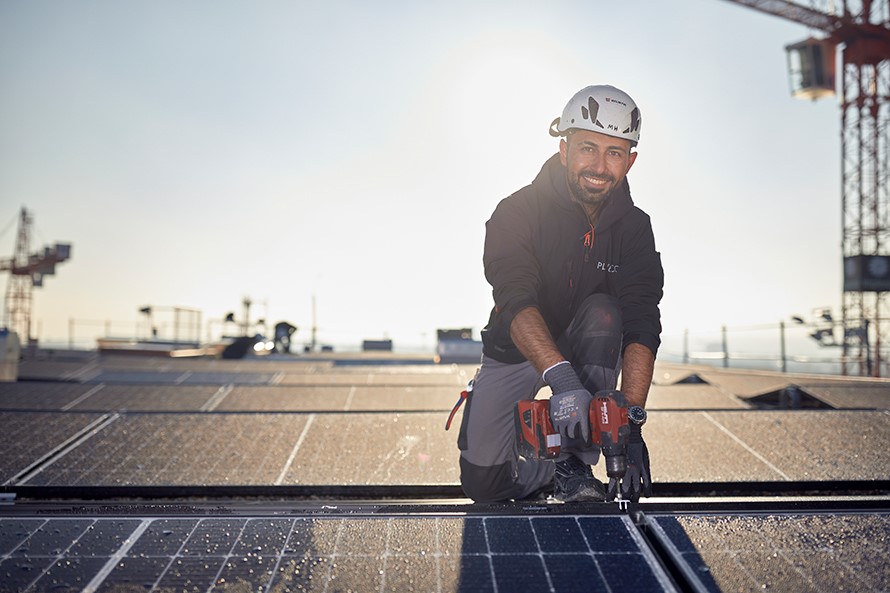 Ein Mann kniet auf einem Flachdach und hat einen Akkuschrauber in der Hand, mit dem er Solarpanels montiert.