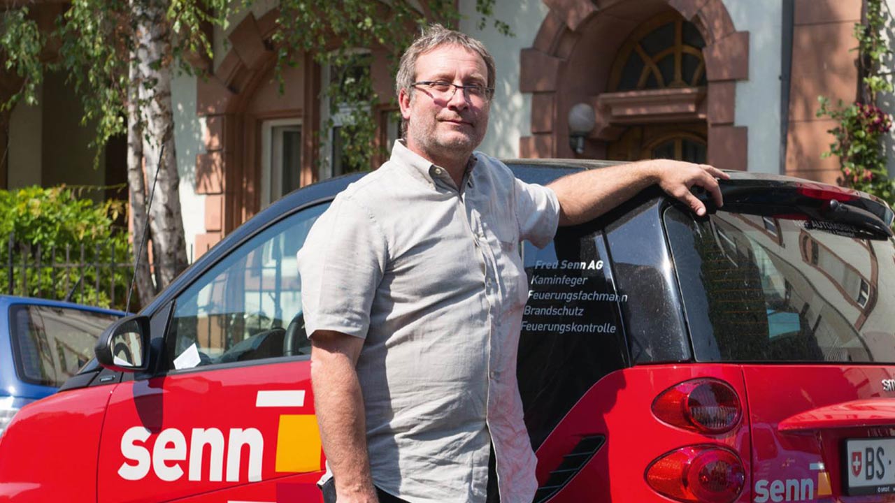 Ein Mann steht vor einem kleinen Auto in einer stästischen Strasse.