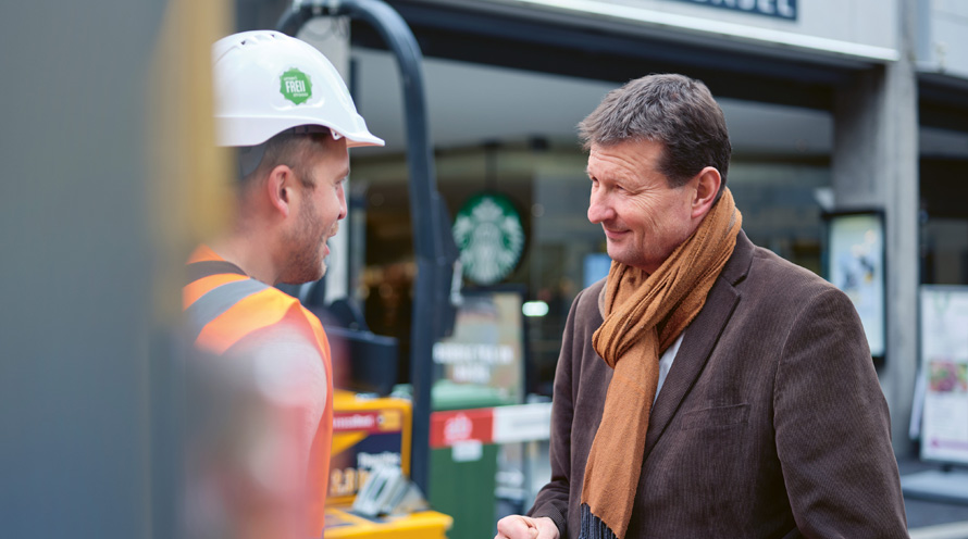 Zwei Männer im Gespräch auf einer Baustelle. Der eine trägt einen Helm, der andere einen Wintermantel mit Schal.