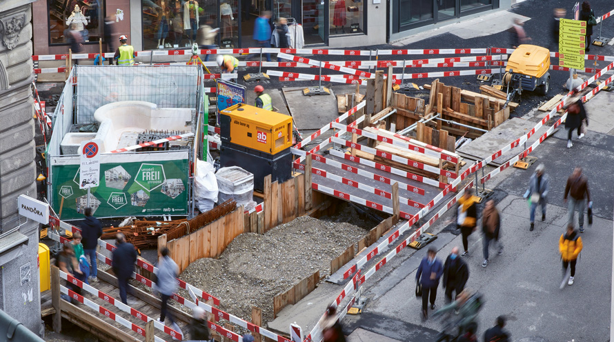 Blick von oben auf eine Baustelle, neben der Menschen die Strasse entlanglaufen.