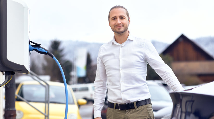 Ein Mann steht zwischen einer Ladestation für E-Autos und einem E-Auto. Er schaut lächelnd in die Kamera.