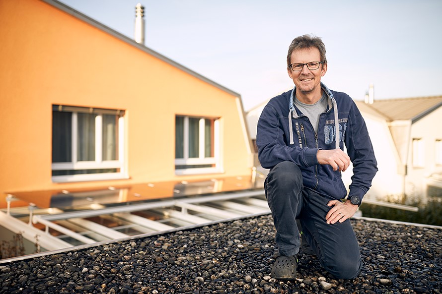 Ein Mann kniet auf einem Flachdach. Im Hintergrund ist das Obergeschoss eines Einfamilienhauses zu sehen, das auf dem Wintergarten eine kleine Solaranlage hat.