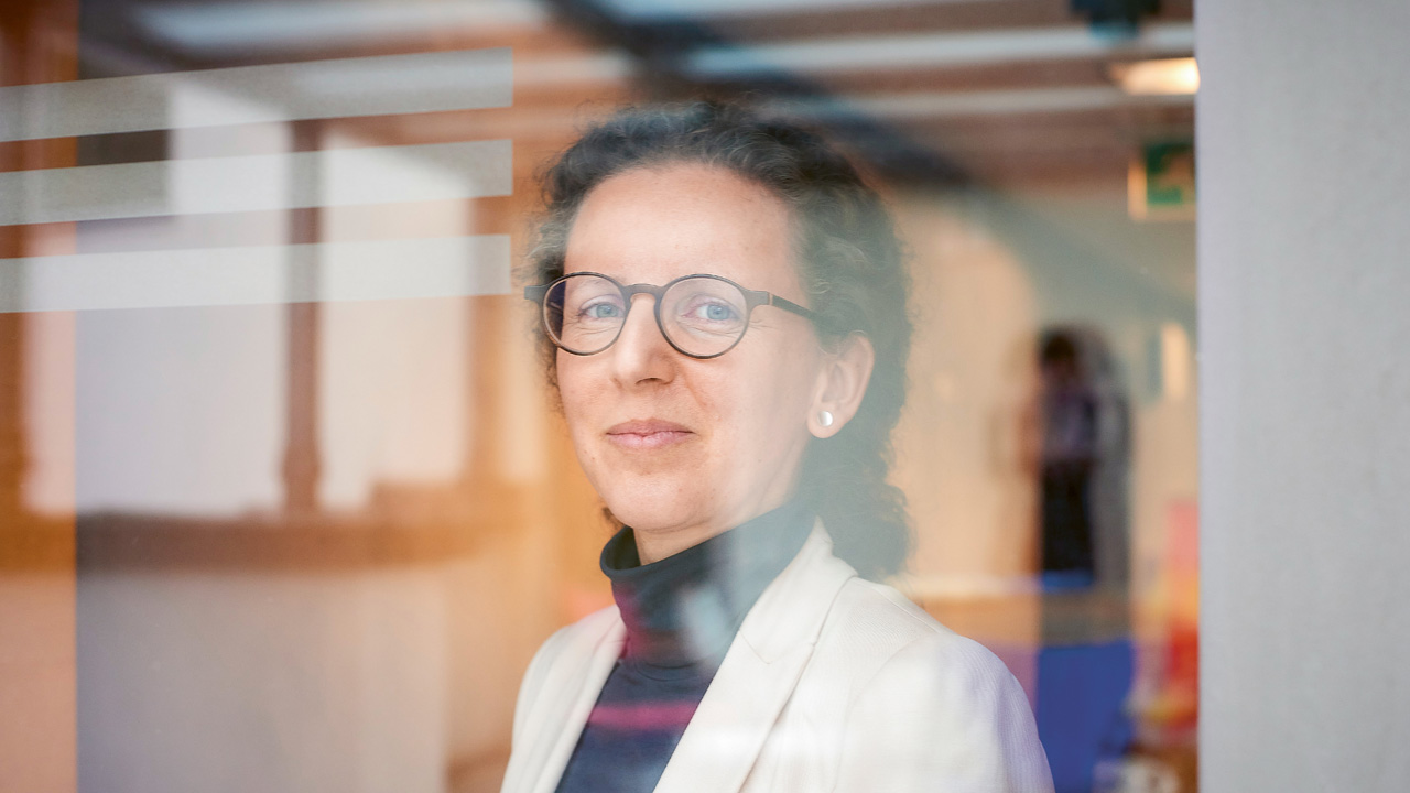 Eine Frau mit Brille hinter einer Glasscheibe schaut in die Kamera.