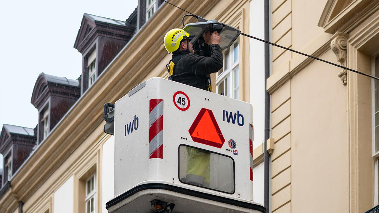 Mann mit Helm steht in einer Arbeitsbühne und hantiert an einer Leuchte der öffentlichen Beleuchtung.