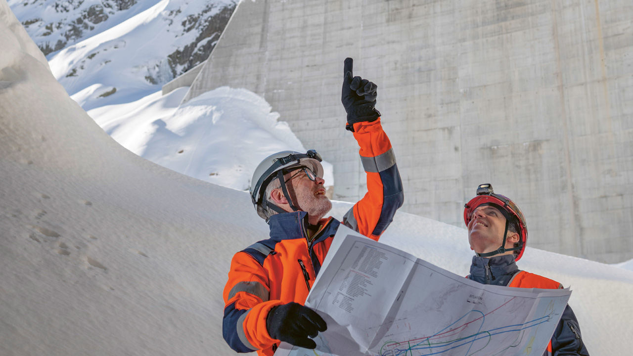 Zwei Männer mit Helm stehen draussen im Schnee. Einer hält eine Karte in der Hand und zeigt in die Luft. Im Hintergrund sieht man eine Staumauer.