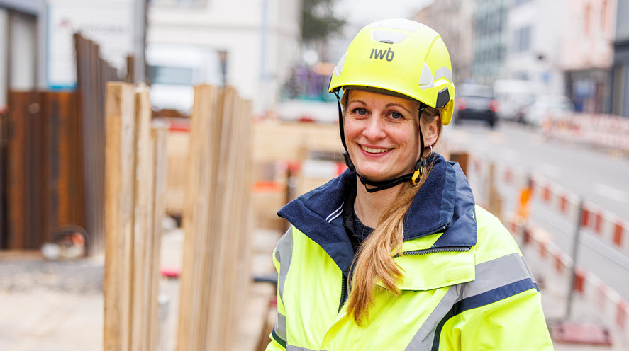 Frau trägt Helm und Leuchtweste und sie steht vor einer Baustelle. 