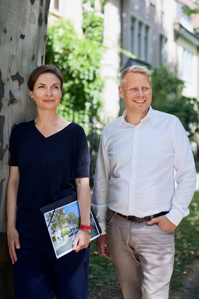 Friederike Meinhardt und Steffen Stoll stehen im Bachlettenquartier neben einer Grünflächen. Sie besprechen den Fernwärmeausbau und die städtebauliche Begrünung.