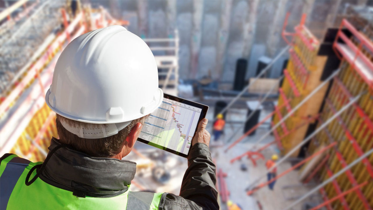 Mann mit Helm schaut auf Tablet. Dort sieht man die Planung eines Projekts. Im Hintergrund ist die Baugrube einer grossen Baustelle.