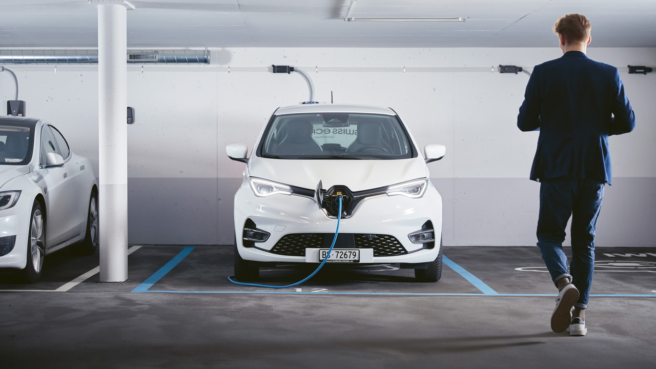 Ein Mann geht auf ein E-Auto zu, das in einer Einstellhalle steht und mit dem Kabel an eine Ladesäule angeschlossen ist.