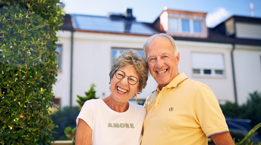 Eine Frau und ein Mann stehen Arm in Arm vor einem Reihenhaus. Auf dessen Dach sind Solarpanels zu sehen.