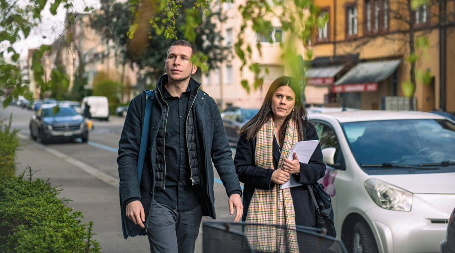 Philipp Frey und Ingela Lakatos in einem Wohnquartier zu Fuss unterwegs. Im Hintergrund sind parkierte Autos zu sehen.