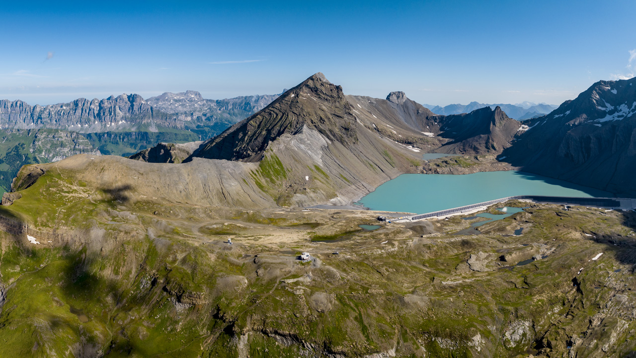 Blick aus der Vogelperspektive auf eine Berglandschaft. In der rechten Bildhälfte ist ein Stausee zu sehen, auf dessen Staumauer eine Photovoltaikanlage ist.