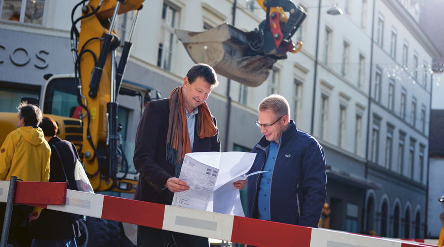 Zwei Männer stehen vor einer Baustelle in der Stadt. Der Linke hält einen Plan in der Hand, im Hintergrund ist eine Baggerschaufel zu sehen.