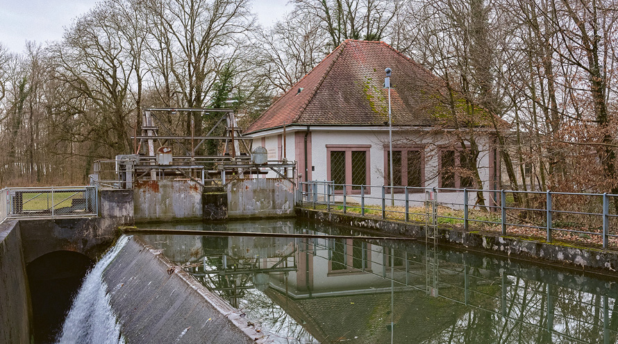 Blick auf das Kraftwerk mit Stauwehr und Becken. Im Hintergrund sieht man das Kraftwerk-Gebäude.