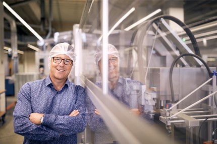 Ein Mann steht neben einer Fabrikationsanlage, die durch eine Plexiglasscheibe von ihm getrennt ist.