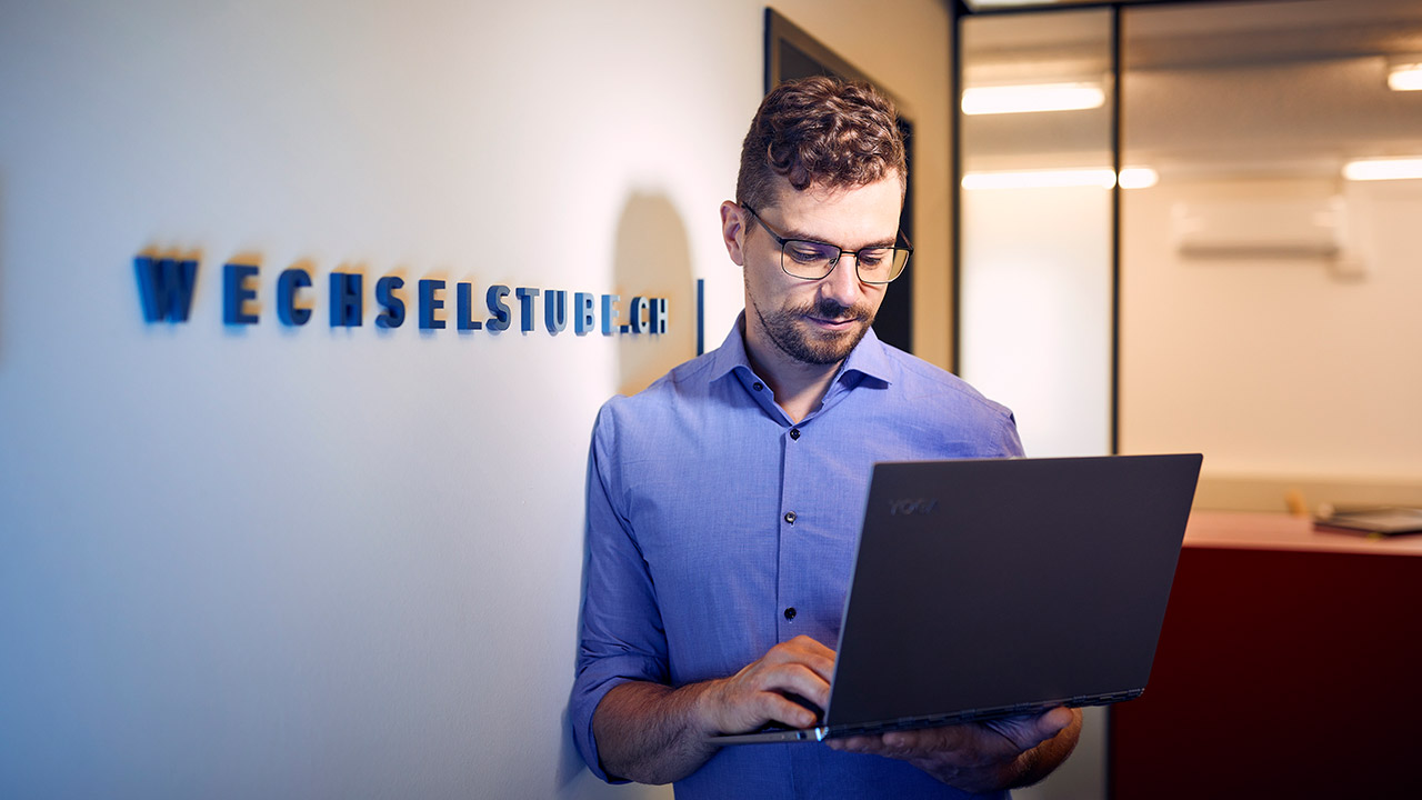 Ein Mann steht mit einem Laptop in einem Büro und lehnt an der Wand.