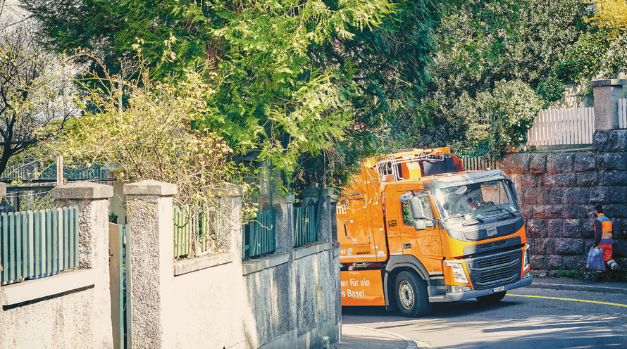 Ein elektrischer Kehrichtlastwagen fährt eine Quartierstrasse hinauf. Ein Belader trägt einen Kehrichtsack.