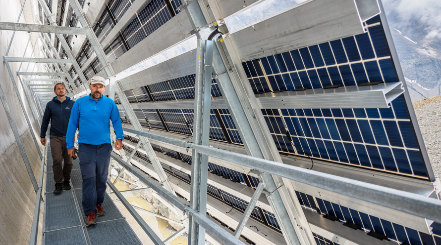 Zwei Männer laufen auf einem Metallsteg hinter einer Solaranlage hindurch. 