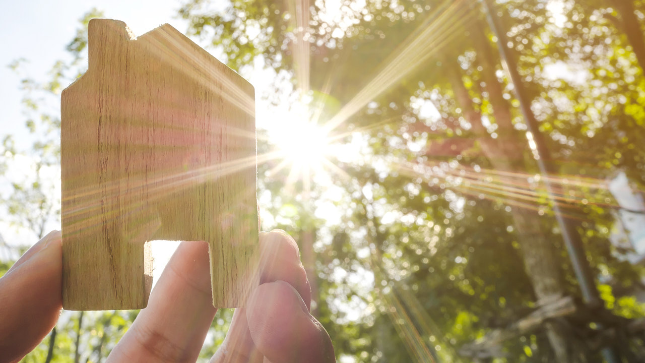 Eine Hand hält ein kleines Holzstück, das wie ein Haus geformt ist, gegen die Sonne.