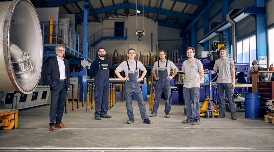 Fünf Männer stehen in einer Fabrikhalle. Ganz links in Anzug steht der Verwaltungsratspräsident, rechts von ihm seine Mitarbeiter.