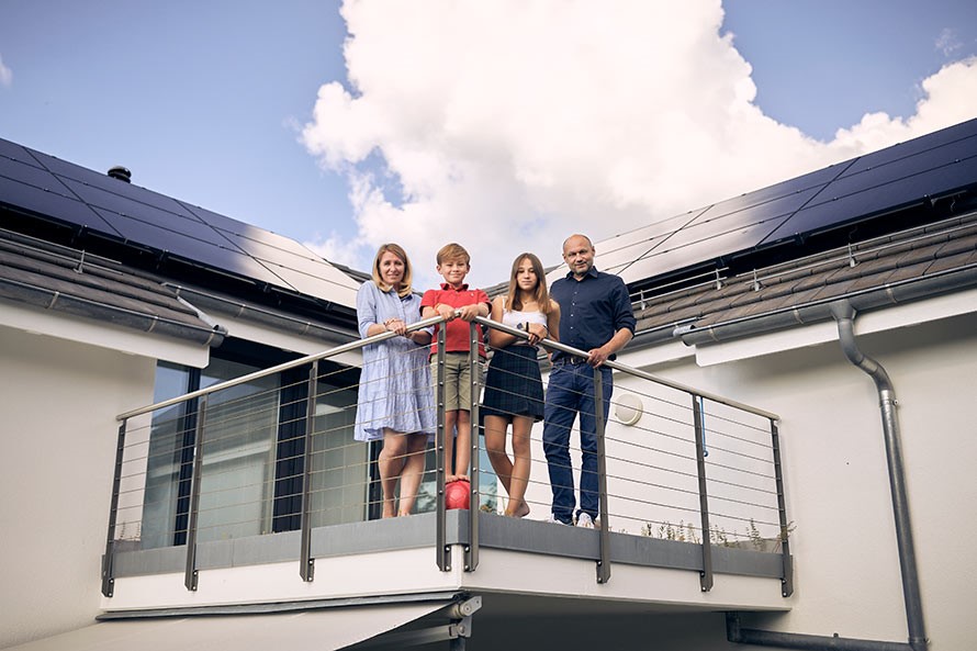 Eine Familie mit Mutter, Vater, Tochter und Sohn steht auf dem Balkon eines Einfamilienhauses. Auf dem Dach im Hintergrund befinden sich Solarpanels.