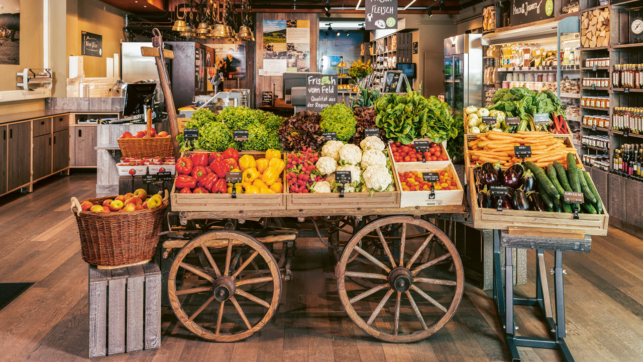 Eine Holzkutsche steht in einem Hofladen. Darauf sind Salate, Peperoni, Blumenkohl, Tomaten und anderes Gemüse.