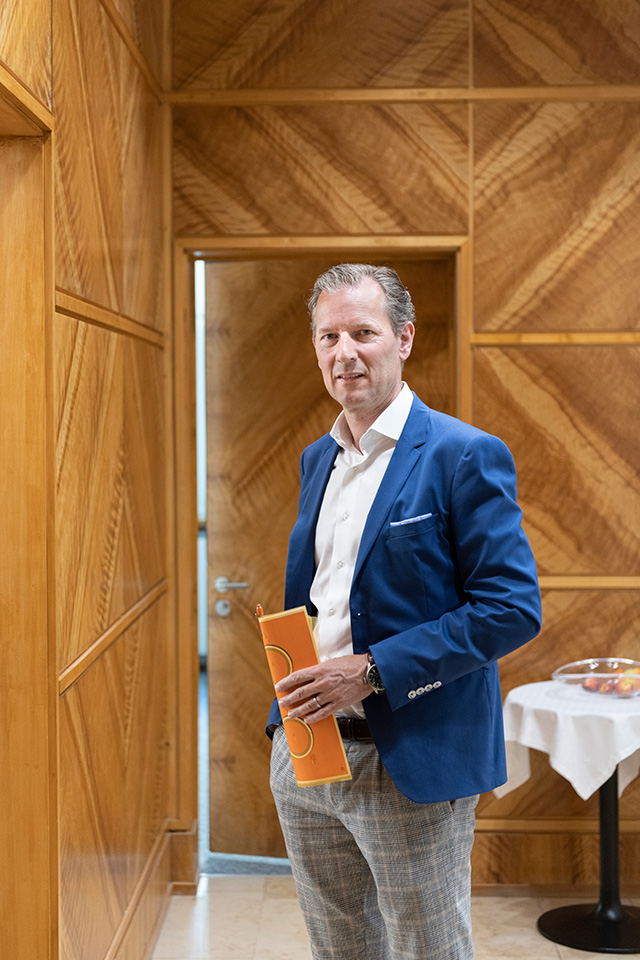 Daniel Petitjean von Warteck Invest AG steht in einem mit Holz ausgestatteten Raum. Er hält eine orange Mappe in der Hand.