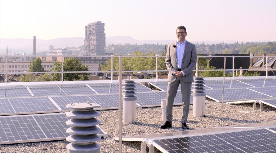 Ein Mann steht auf einem Flachdach zwischen Solarpanels. Im Hintergrund ist ein Hochhaus zu sehen.