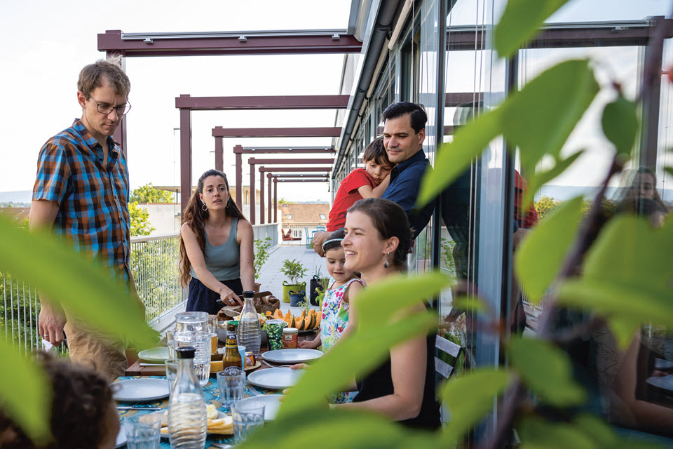 Menschen und Kinder sitzen am und stehen rund um einen Tisch auf der Terrasse. Der Tisch ist gedeckt.